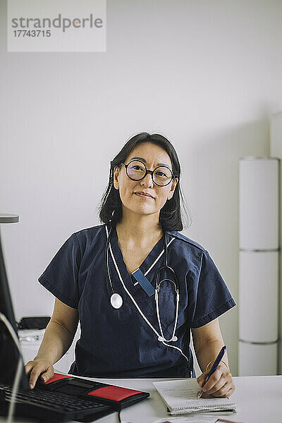 Porträt einer lächelnden Ärztin mit Brille  die am Schreibtisch in einer medizinischen Klinik sitzt und schreibt