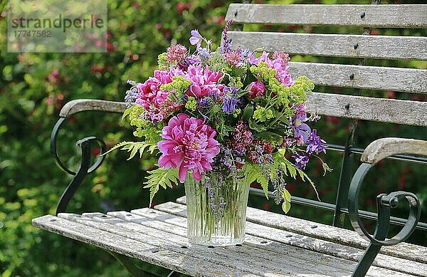 Bunter Blumenstrauß in rot  pink und violett Farbtönen mit Pfingstrosen und Akeleien  steht in Glas-Vase auf dekorativer Holz-GartenbankBunter Blumenstrauß in rot  steht in Glas-Vase auf dekorativen Holz-Gartenbank