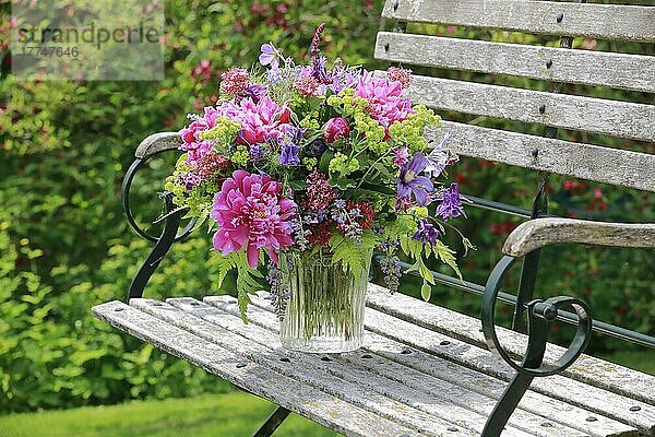 Bunter Blumenstrauß in rot  pink und violett Farbtönen mit Pfingstrosen und Akeleien  steht in Glas-Vase auf dekorativer Holz-Gartenbank
