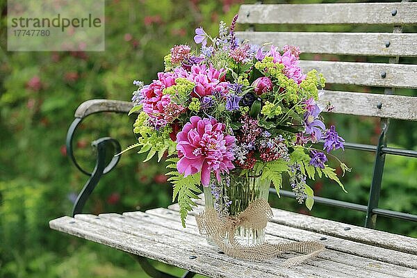 Bunter Blumenstrauss in rot  pink und violett Farbtönen mit Pfingstrosen und Akeleien  steht in Glas-Vase auf dekorativen Holz-Gartenbank