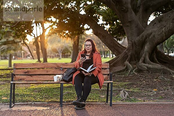 Eine leitende Frau in Übergröße arbeitet im Freien. Sie hält ein Notebook und sendet eine Textnachricht  während sie auf einer Bank in einem öffentlichen Park sitzt