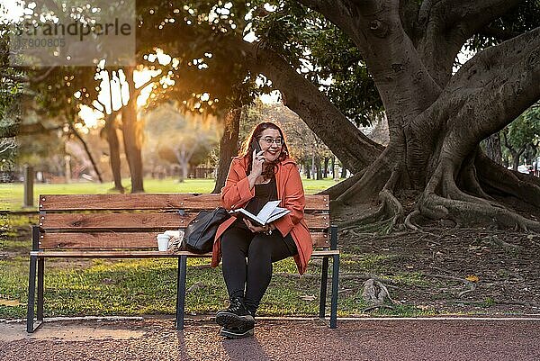 Eine leitende Frau in Übergröße arbeitet im Freien. Sie hält ein Notebook und führt ein Telefongespräch  während sie lacht und auf einer Bank in einem öffentlichen Park sitzt