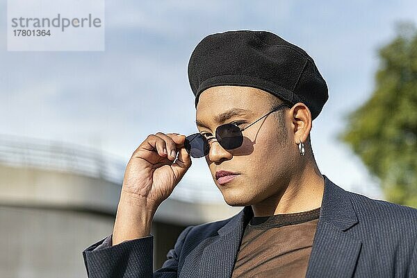 Young latin gay man with make-up on wearing a fashionable hat and looking over sunglasses at camera. LGBT