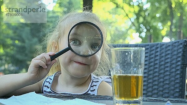 Kleines Mädchen schaut in die Linse um. Nahaufnahme eines blonden Mädchens  das die Welt um sich herum studiert und sie durch ein Vergrößerungsglas betrachtet  während es in einem Straßencafé im Park sitzt. Odessa  Ukraine  Europa