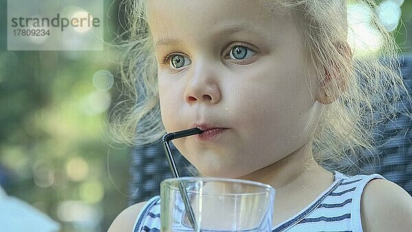 Süßes kleines Mädchen trinkt Saft durch Strohhalm. Close-up-Porträt von blonden Mädchen trinkt Saft aus Glas durch Cocktail Stroh sitzen in Straßencafé auf dem Park. Odessa  Ukraine  Europa