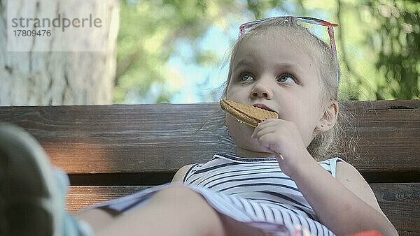 Kleines Mädchen isst Lebkuchen. Close-up-Porträt von blonden Mädchen sitzen auf Parkbank und essen Kekse. Odessa Ukraine