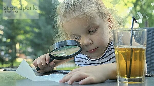 Kleines Mädchen schaut vorsichtig in die Linse auf das Salz. Nahaufnahme eines blonden Mädchens  das Salzkristalle studiert  während es sie durch ein Vergrößerungsglas betrachtet  während es im Straßencafé im Park sitzt. Odessa  Ukraine  Europa