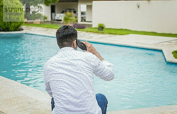 Hübscher Mann sitzt und telefoniert in der Nähe des Swimmingpools  Junger Mann sitzt und telefoniert am Swimmingpool  Mann sitzt und telefoniert am Rande des Swimmingpools