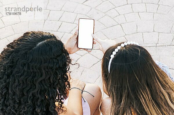 Hände von zwei Mädchen zeigen auf Handy-Bildschirm  Zwei Mädchen sitzen und zeigen auf leeren Handy-Bildschirm mit Platz für Text