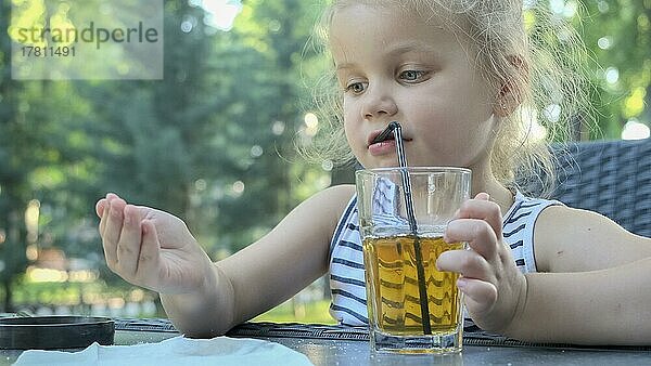 Süßes kleines Mädchen trinkt Saft durch Strohhalm. Close-up-Porträt von blonden Mädchen trinkt Saft aus Glas durch Cocktail Stroh sitzen in Straßencafé auf dem Park. Odessa  Ukraine  Europa