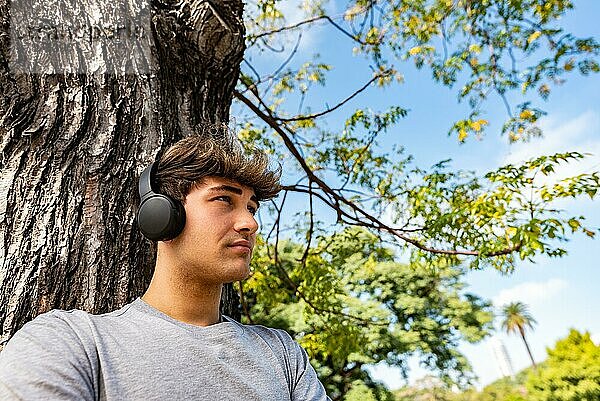 Ein junger Mann  der Musik hört und sich mit verschränkten Armen an einen Baum lehnt
