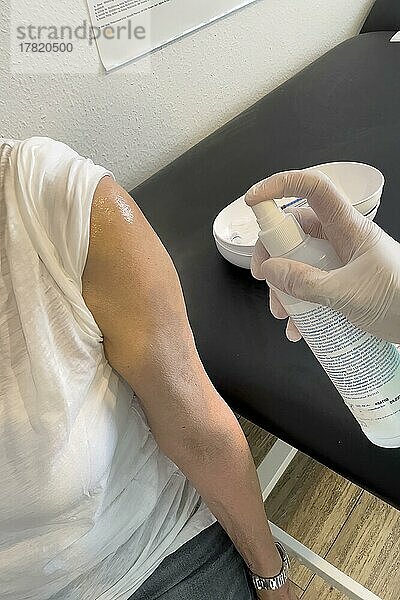 Arm von Patientin wird von Arzthelferin desinfiziert mit medizinsches Desinfektionsmittel Spray Oberarm vor Injektion für Impfung Booster gegen Covid-19 Corona-Viren Corona-Virus