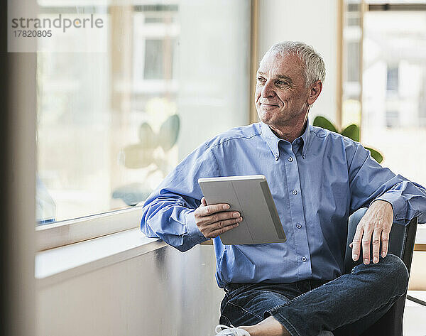 Smiling senior businessman with tablet PC sitting by window