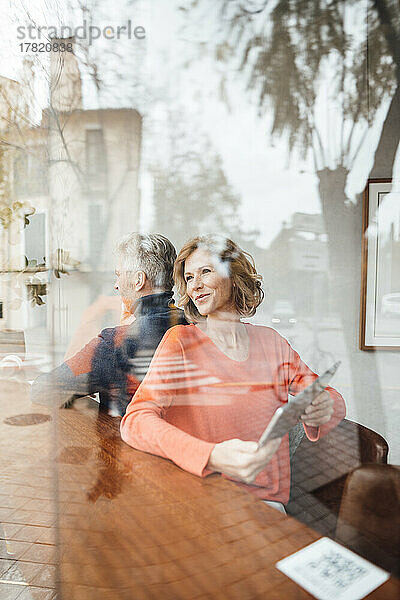 Lächelnde Frau mit Tablet-PC sitzt neben Mann im Café und sieht durchs Fenster