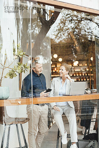 Lächelnde Geschäftsfrau mit Geschäftsmann  der sein Mobiltelefon im Café hält und durch die Glasscheibe gesehen wird