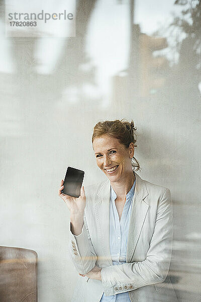Glückliche Geschäftsfrau zeigt Mobiltelefon im Café  gesehen durch Glas