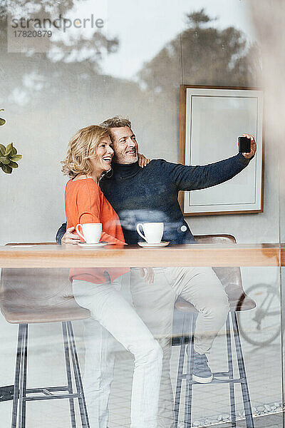 Lächelnder Mann und Frau machen Selfie im Café  gesehen durch Glasfenster