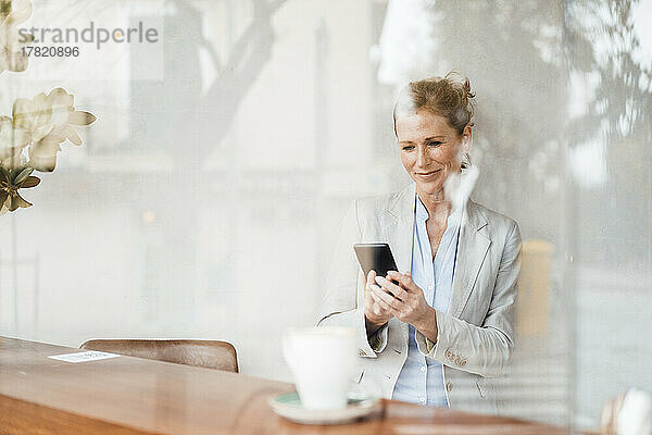 Lächelnde Geschäftsfrau blickt durch Glas auf ihr Smartphone im Café