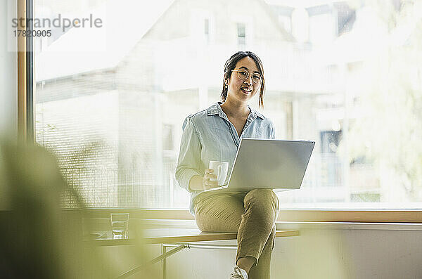 Lächelnde Geschäftsfrau mit Laptop und Kaffeetasse am Schreibtisch