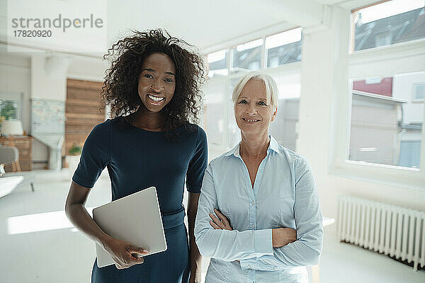 Smiling businesswoman holding laptop standing with colleague in office