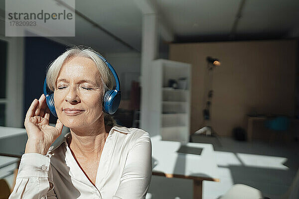 Lächelnde ältere Frau mit geschlossenen Augen  die im Büro Musik über drahtlose Kopfhörer hört