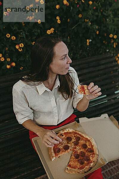 Frau isst ein Stück Pizza und sitzt auf einer Bank