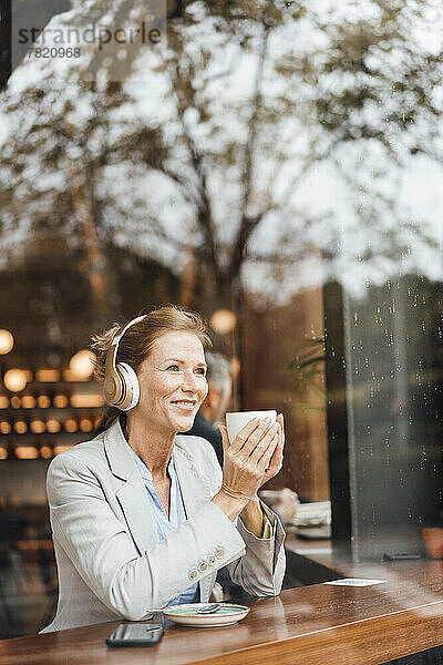 Lächelnde Geschäftsfrau hält Kaffeetasse in der Hand und hört Musik im Café  gesehen durch Glas