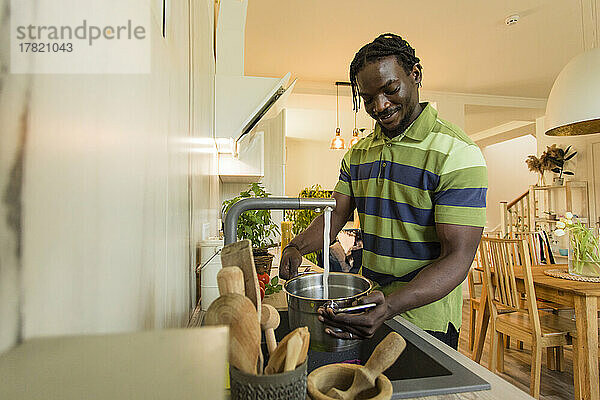 Lächelnder junger Mann füllt Topf aus Wasserhahn in Küche