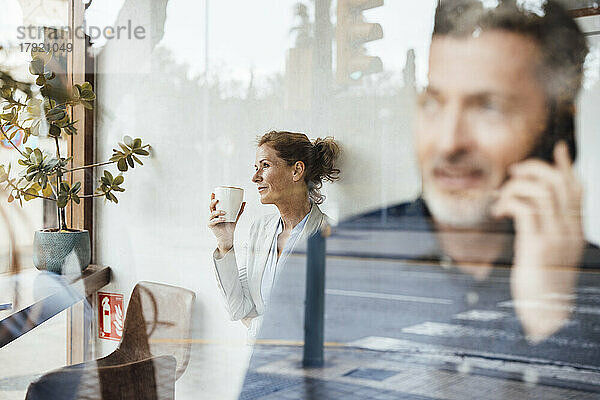 Ein Geschäftsmann telefoniert mit einer Geschäftsfrau  die im Hintergrund eine Kaffeetasse hält und durch Glas gesehen wird