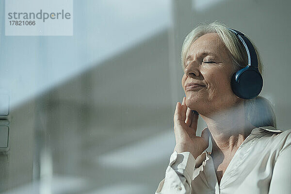 Lächelnde ältere Frau mit geschlossenen Augen  die über drahtlose Kopfhörer Musik hört