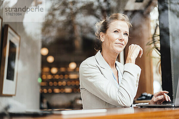 Lächelnde Geschäftsfrau sitzt mit Laptop im Café und sieht durch Glas