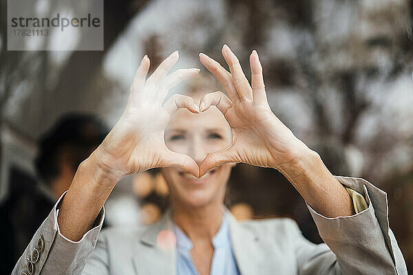 Lächelnde Geschäftsfrau gestikuliert Herzform im Café  durch Glas gesehen