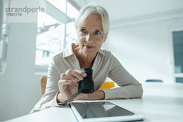 Ältere Geschäftsfrau hält Lupe und sitzt mit Tablet-PC im Büro