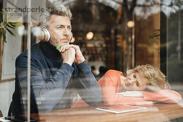 Man listening music through wireless headphones sitting by woman sleeping on table in cafe