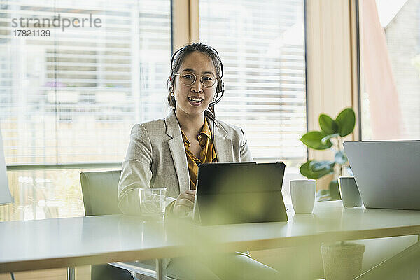 Glückliche Geschäftsfrau mit Headset sitzt am Schreibtisch im Büro