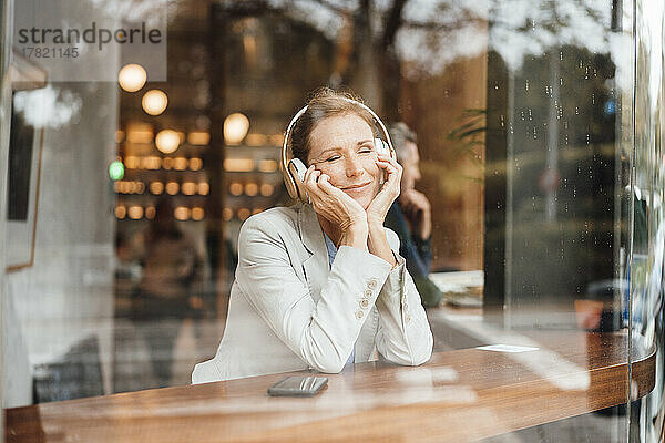 Lächelnde Geschäftsfrau mit geschlossenen Augen  die im Café Musik hört und durch Glas gesehen wird