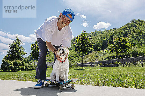 Glücklicher Mann mit Hund sitzt auf Skateboard im Park
