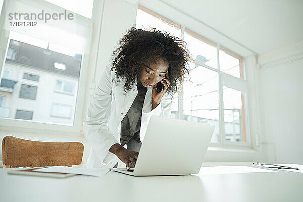 Female doctor talking on mobile phone using laptop at desk