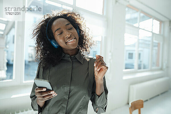Junge Frau hält ihr Mobiltelefon in der Hand und genießt Musik  die sie im Büro über drahtlose Kopfhörer hört