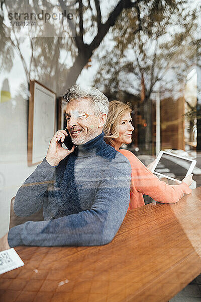 Lächelnder Mann  der im Café mit einem Smartphone spricht  während eine Frau einen Tablet-PC in der Hand hält