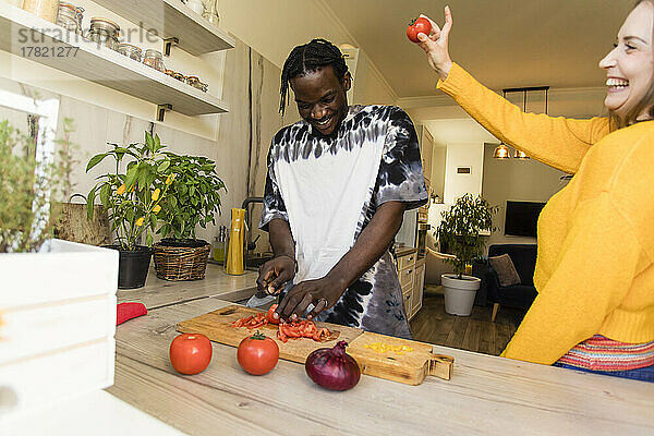 Glückliche Frau hält Tomate und steht neben ihrem Freund  der in der Küche Gemüse schneidet