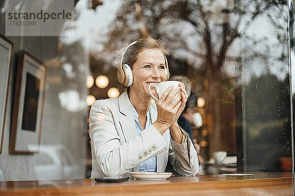 Lächelnde Geschäftsfrau hält Kaffeetasse in der Hand und hört im Café Musik über kabellose Kopfhörer