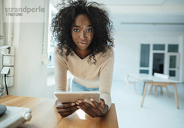 Geschäftsfrau mit lockigem Haar hält Tablet-PC in der Hand und stützt sich auf den Schreibtisch im Büro
