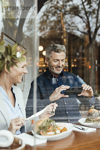 Lächelnder Geschäftsmann fotografiert Mahlzeit per Smartphone von Geschäftsfrau im Café