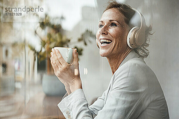 Glückliche Geschäftsfrau hält Kaffeetasse in der Hand und hört Musik über drahtlose Kopfhörer im Café