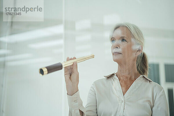 Ältere Geschäftsfrau hält Teleskop im Büro  gesehen durch Glas
