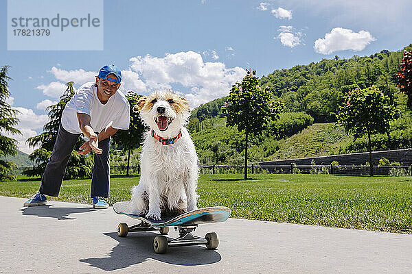 Hund fährt Skateboard  Mann steht dahinter im Park