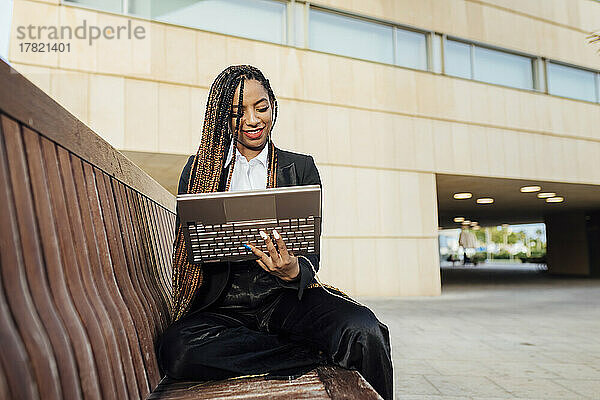 Glückliche Geschäftsfrau sitzt mit Laptop auf Bank vor Bürogebäude