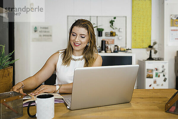 Lächelnde junge Geschäftsfrau mit Smartphone und Laptop sitzt am Schreibtisch im Büro
