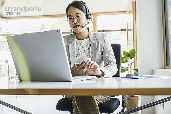 Lächelnde Geschäftsfrau mit Headset  die am Schreibtisch am Laptop arbeitet